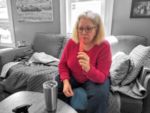 Heather sitting on a couch with a single stick popsicle near her mouth. The photo is in greyscale except for Heather who is in a full vibrant colour with a pink sweater and blue jeans.