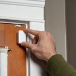 The upper corner of a wooden door, with a hand holding one half of a two part electronic door sensor, presumably installing it.